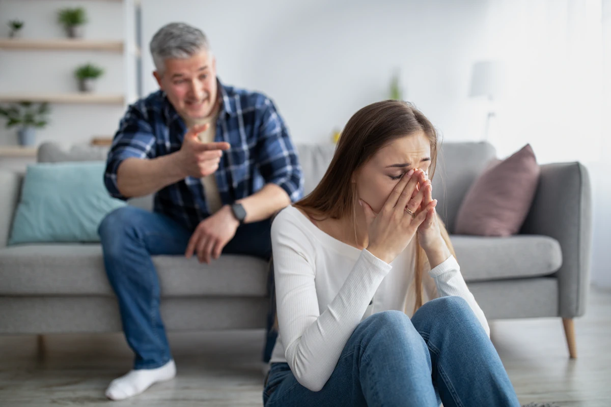 Une femme qui pleure assise par terre et un homme à l'arrière qui continue de lui pointer doigt