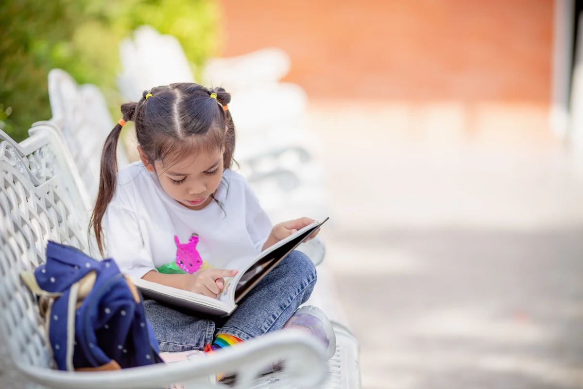 Une petite fille assise sur un banc à l'extérieur en train de lire un livre