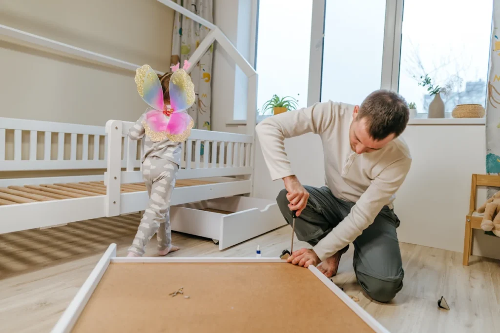 Un homme qui monte un meuble dans une chambre d'enfant avec une petire fille à côté