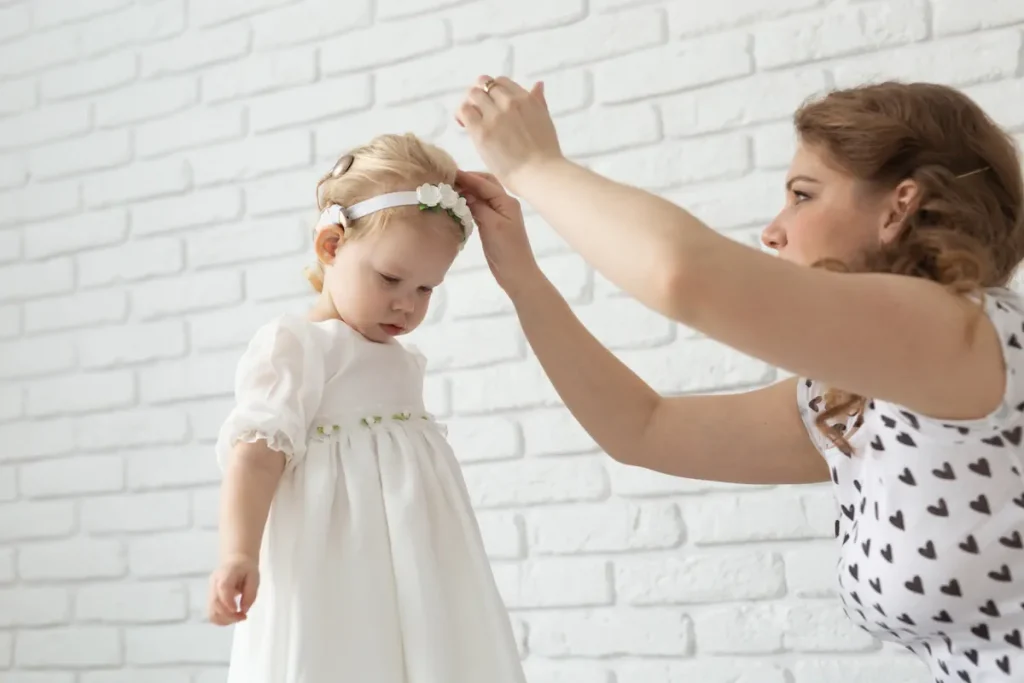 Une maman en robe qui habille sa fille avec une robe blanche et un bandeau blanc